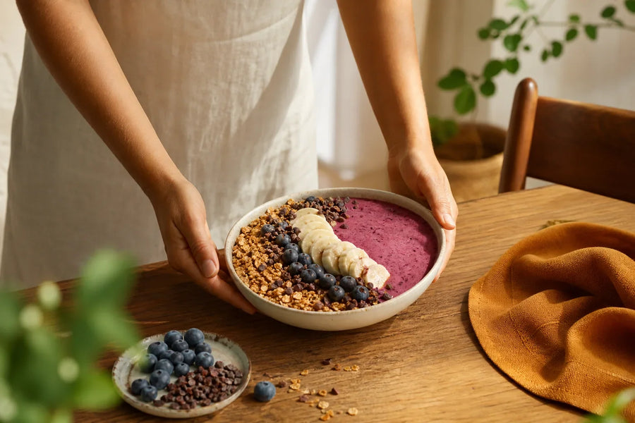 Pessoa apoiando smoothie bowl com banana, blueberries, cacau e granola.